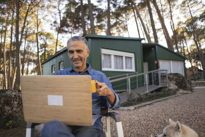 Smiling mature man with cup of coffee sitting outside green building using laptop