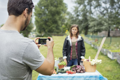 Mid adult man photographing woman standing by freshly harvested vegetables in urban garden