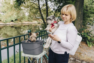 Blond mature woman with cute dogs using smart phone while standing by bicycle in park