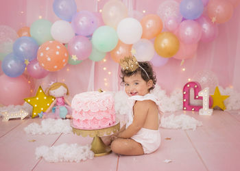 Cute baby girl with balloons and birthday cake sitting at home