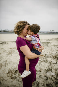 Side view of pregnant mother with son rubbing noses son while standing at beach against sky