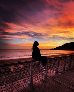 Silhouette woman sitting on railing against sea during sunset