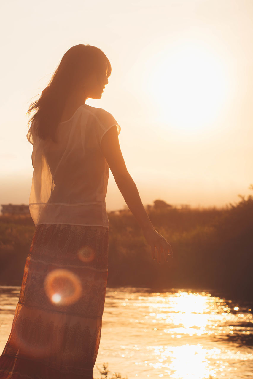 sunset, real people, one person, water, sun, leisure activity, nature, standing, lifestyles, outdoors, beauty in nature, sky, rear view, sunlight, women, childhood, scenics, sea, full length, clear sky, tree, day, young adult, people