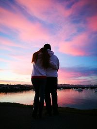 Rear view of couple standing on shore against sky during sunset