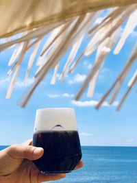 Hand holding glass of water at beach against sky