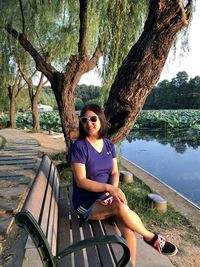 Portrait of woman wearing sunglasses sitting on bench against trees