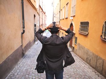 Rear view of man standing with hands behind head at aisle amidst buildings