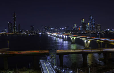 Illuminated bridge over river by buildings against sky at night