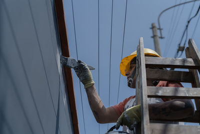 From below professional male painter in yellow hardhat in process of plastering wall while standing on a ladder on sidewalk on city street