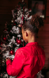 Back view of cheerful little black girl in red shirt looking at camera and smiling while standing near decorated christmas tree at home