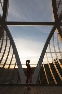 Side view of woman standing on footbridge against sky at sunset