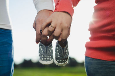 Cropped hands of couple holding baby booties against sky