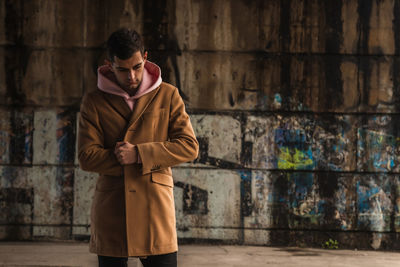 Young man wearing trench coat while standing against wall outdoors