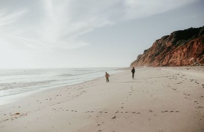 Siblings walking at beach against sky during sunny day