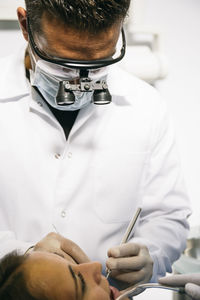 Male dentist in surgical loupes and mask examining teeth of female patient in clinic