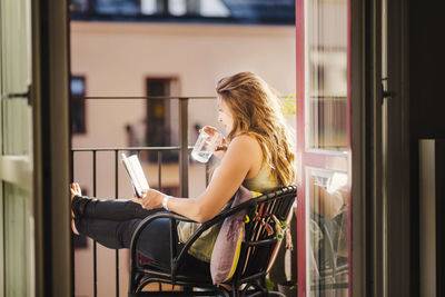 Side view of woman drinking water while reading guidebook at balcony