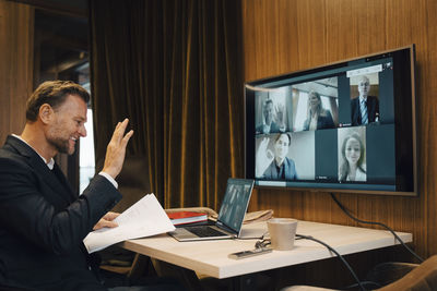 Smiling businessman waving with colleagues on video call during meeting in boardroom