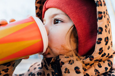 Side view of cute girl in warm clothes drinking takeaway hot beverage from paper cup while standing on street on cold day