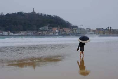 A woman with an umbrella walks along the beach shore overseeing the san sebastian, spain