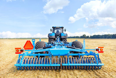 A trailed blue harrow on agricultural tractor against the backdrop of harvested yellow wheat field 