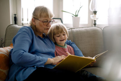 Grandmother reading storybook for grandson while sitting on sofa in living room