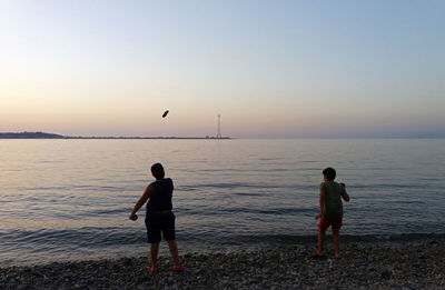 Rear view of men standing at beach against sky during sunset