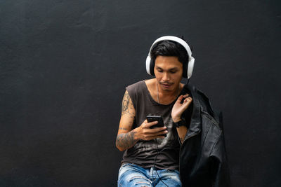Man wearing headphones while using mobile phone against wall