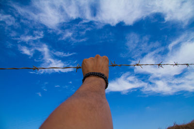 Cropped hand of man holding barbed wire against blue sky