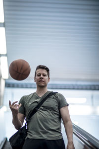 Low angle portrait of young man playing with basketball while moving down on escalator