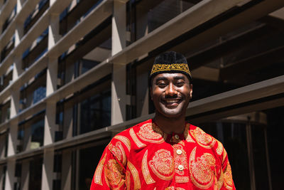 Happy adult african guy in traditional red clothes and kufi cap looking at camera in city street near construction in sunny day