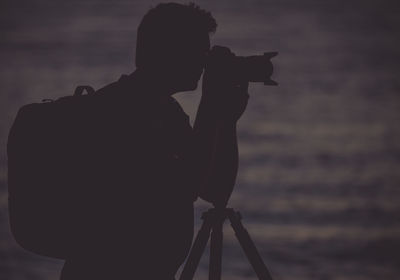 Silhouette photographer carrying backpack standing against sea