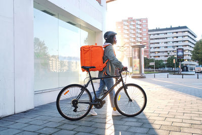 Side view of serious african american female in helmet with orange thermo backpack and smartphone walking with bicycle on road while delivering order