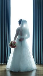 Rear view of bride wearing wedding dress standing with bouquet on hardwood floor