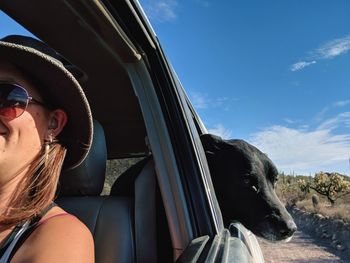 Woman taking selfie of dog looking out of window in car against sky