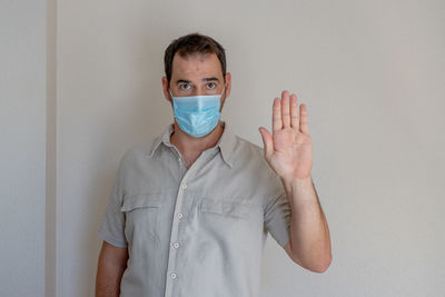 Portrait of man wearing flu mask gesturing while standing against white wall