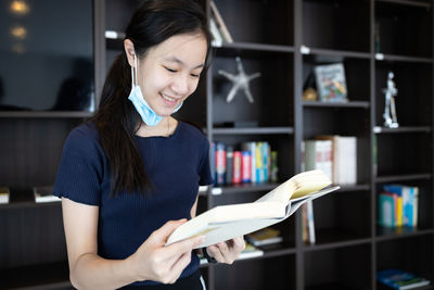 Young woman holding book while standing on shelf