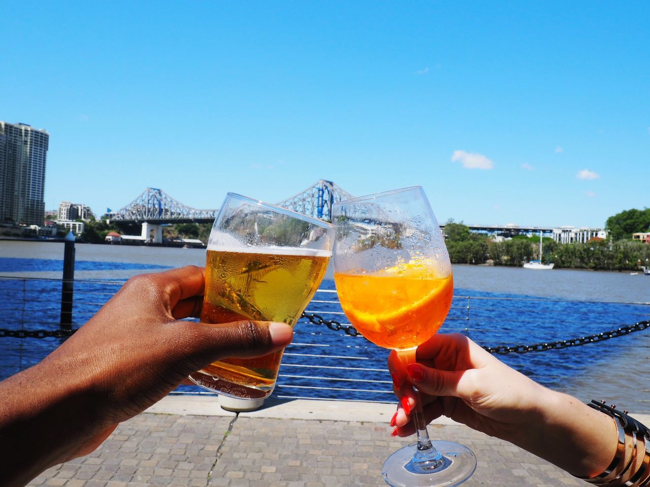 human hand, alcohol, drink, two people, human body part, clear sky, holding, sky, refreshment, food and drink, drinking glass, celebratory toast, water, celebration, real people, outdoors, day, leisure activity, cocktail, close-up, sea, men, architecture, people