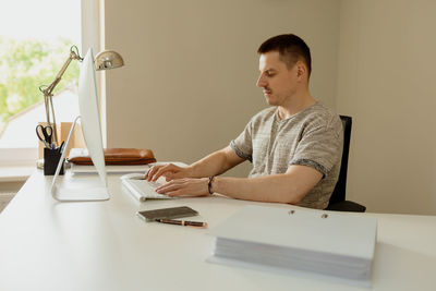Young caucasian man sitting at his desk in the office and working with computer, answering emails. 