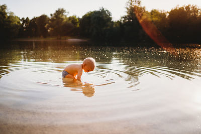 Male toddler looking for stones underwater at lake with reflection