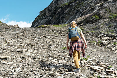 Rear view young woman with backpack in plaid shirt walking uphill along rocky trail in mountain hike