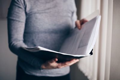 Midsection of businesswoman reading file while standing by window in office