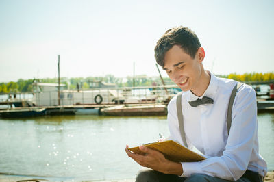 Young man writing in book while sitting at harbor against sky