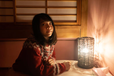 Portrait of young woman sitting by illuminated lamp on table at home