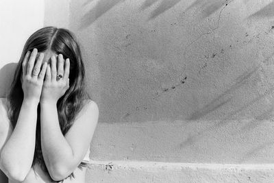 Woman with hands covering face standing against wall