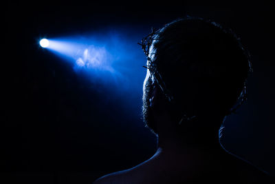 Rear view of man wearing barbed wire against illuminated light in darkroom