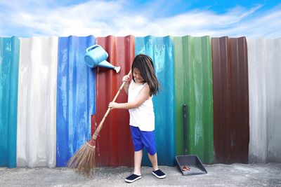 Full length of girl sweeping footpath by colorful corrugated irons