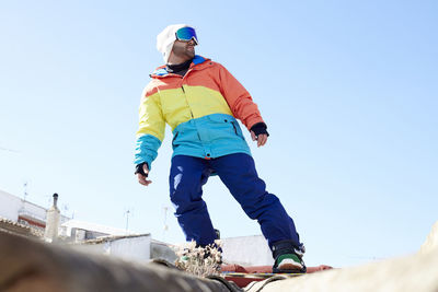 Man snowboarding on a rooftop wearing snow gear and looking at the horizon.