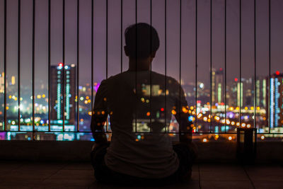Rear view of man sitting against illuminated buildings and sky at night