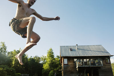 Low angle view of shirtless man jumping into lake against log cabin and clear sky