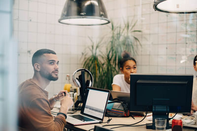 Young male hacker looking away while sitting with laptop at desk in creative office seen through window